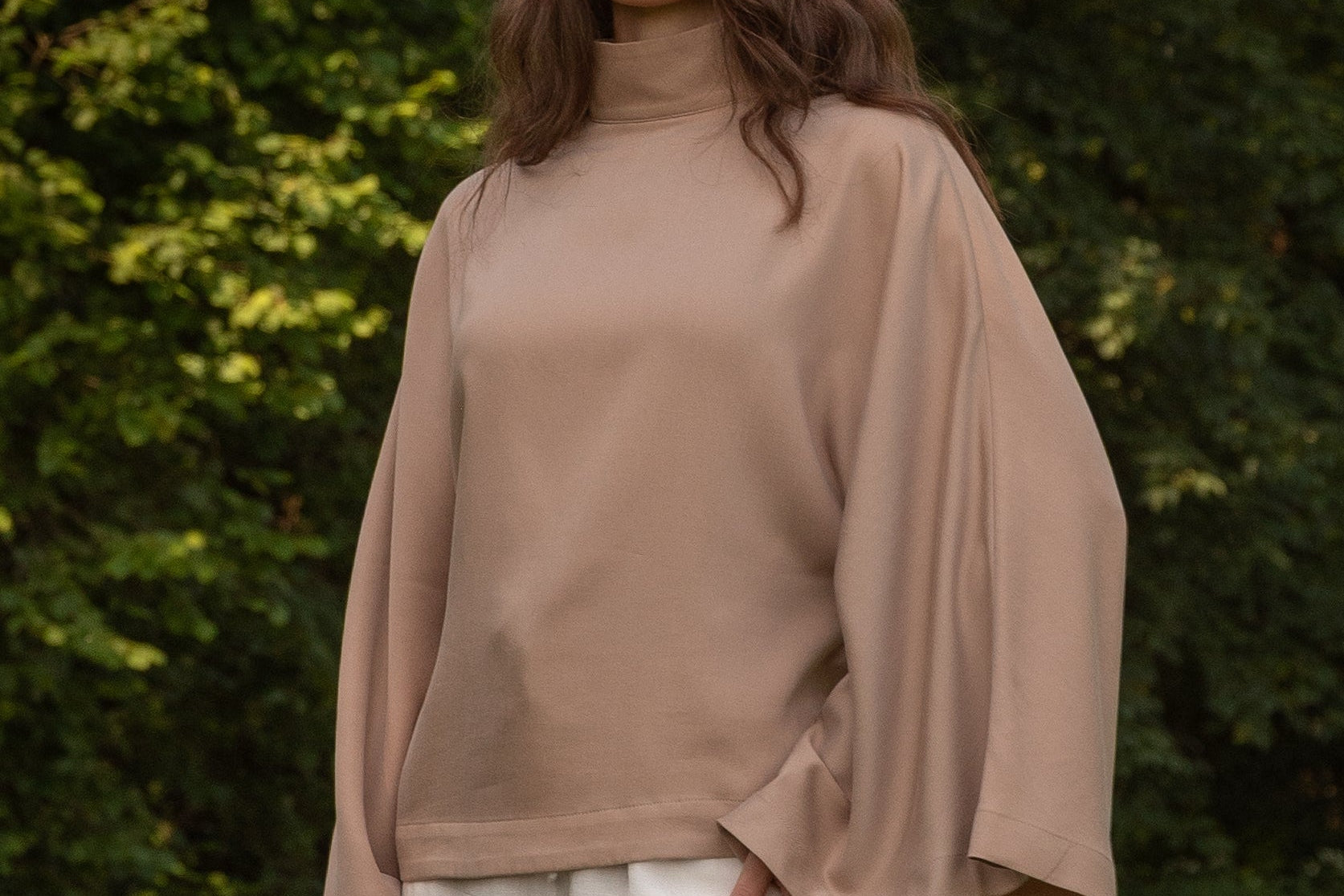 A woman wearing a beige mock-neck long-sleeve top with wide bell sleeves and white pants, standing outdoors in front of a green forest background.